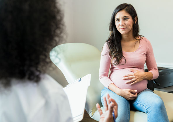 A young woman holds her pregnant belly while she listens to her OB-GYN at a prenatal appointment.