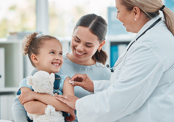 A young girl is smiling, holding a stuffed animal in her mom's lap before getting her flu shot